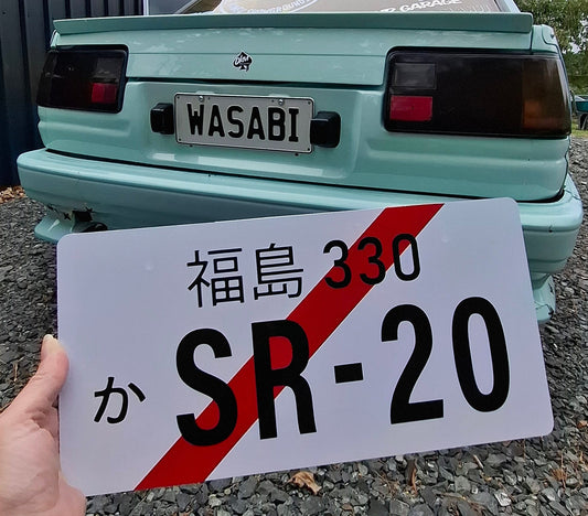 Hand holding a license plate with 'SR-20' on a road next to a vehicle.